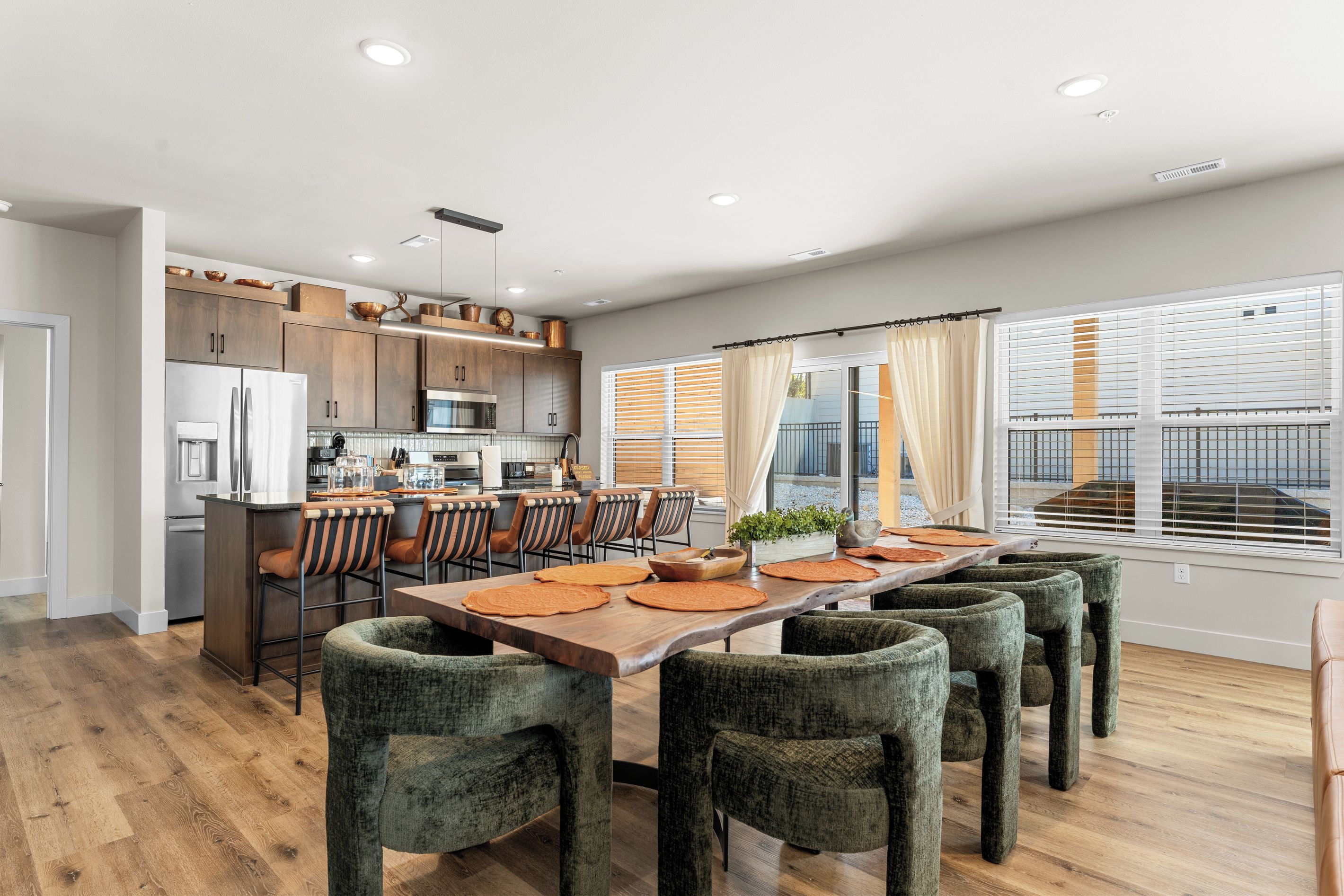 A modern kitchen with stainless steel appliances, a dining area with a long table and chairs, and large windows with blinds.