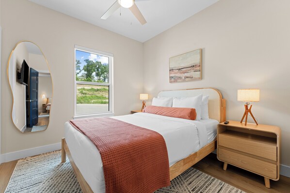 A bedroom with a bed, bedside tables, a ceiling fan, a window with a view, and a painting on the wall. - Taneycomo Wake & Lake | NEW • Private Pool (Branson)