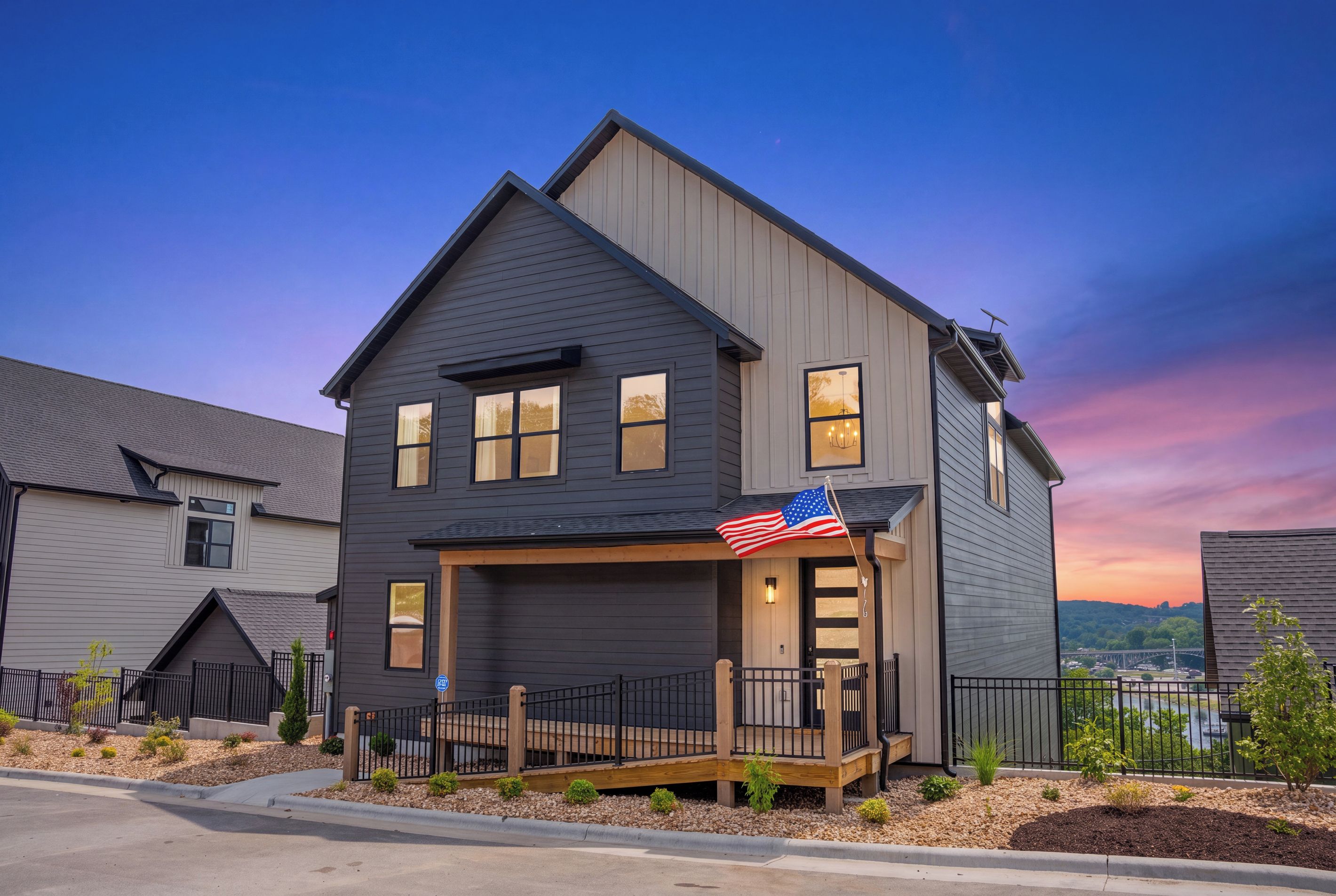 A two-story house with a dark siding exterior, a front porch with a railing, and an American flag.