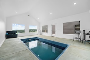 A modern indoor swimming pool with a clear glass bottom, surrounded by white walls and large windows offering views of the outdoors. - Taneycomo Wake & Lake | NEW • Private Pool (Branson)