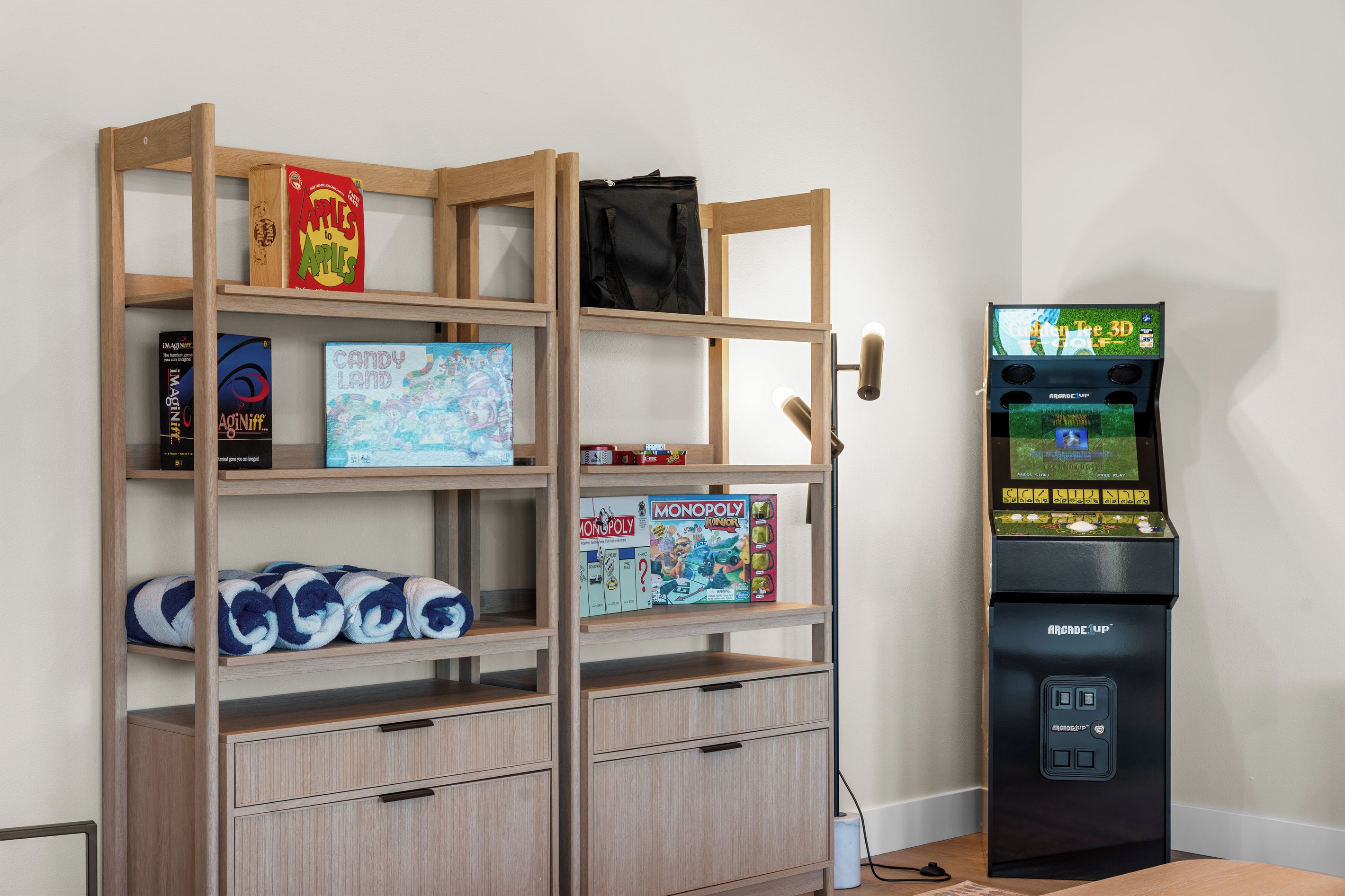 Et rum med en reol i træ, der viser brætspil, et Monopoly Junior-spil og en taske. Der er også en sort arkadespilautomat med skærm og knapper.