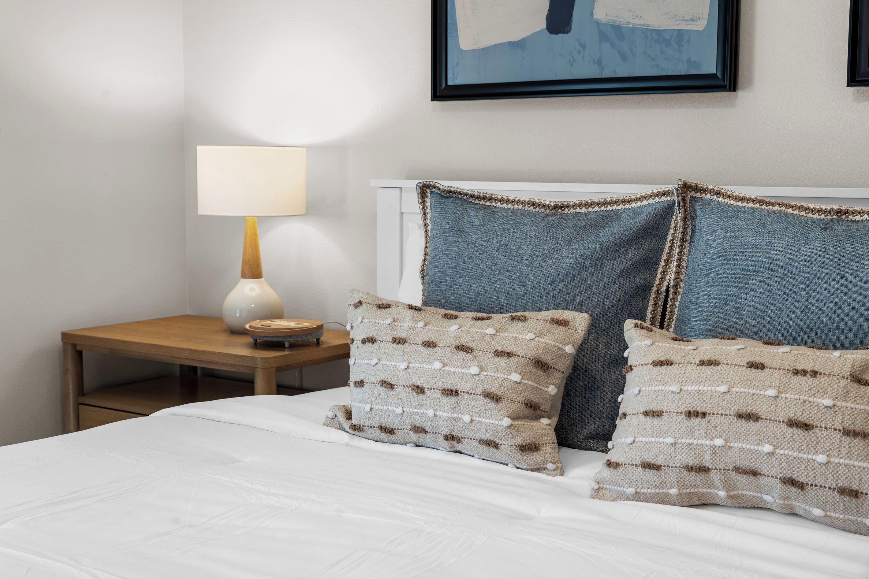 A neatly made bed with white bedding, two decorative pillows with a textured pattern, a bedside table with a lamp, and a framed artwork on the wall.