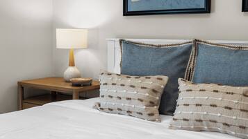 A neatly made bed with white bedding, two decorative pillows with a textured pattern, a bedside table with a lamp, and a framed artwork on the wall.