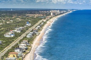 A coastal residential area with beachfront properties, a road parallel to the beach, and distant high-rise buildings. - Sea Turtle Nest | Oceanfront Cottage | Private Beach Access | Dog Friendly | Flagler Beach (Flagler Beach)