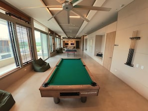 A pool table in a room with large windows, a ceiling fan, and a ping pong table in the background. - SAVIAsamaria. Loft con balcón de vista al mar en lujoso resort (Santa Marta)