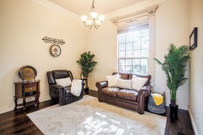 Ein Wohnzimmer mit einem braunen Ledersofa, einem dunklen Holzboden, einem weißen Teppich, einer Kristallleuchtre und einem großen Fenster mit Jalousien.
