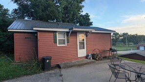 A small, single-story building with a metal roof, a front door, and a window. There is a trash can and a grill outside.