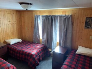 A room with two beds, a wooden wall, a window with curtains, and a ceiling fan.