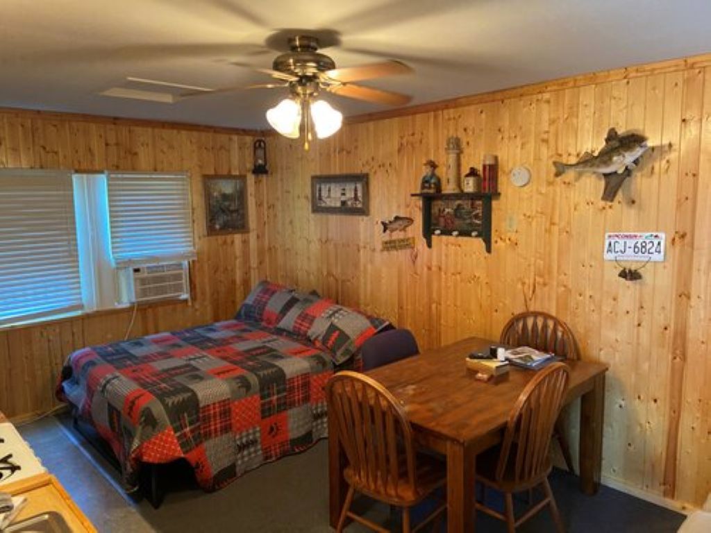 Une pièce avec un mur en bois, un lit recouvert d’une couverture à carreaux, une table à manger avec des chaises et un ventilateur de plafond.