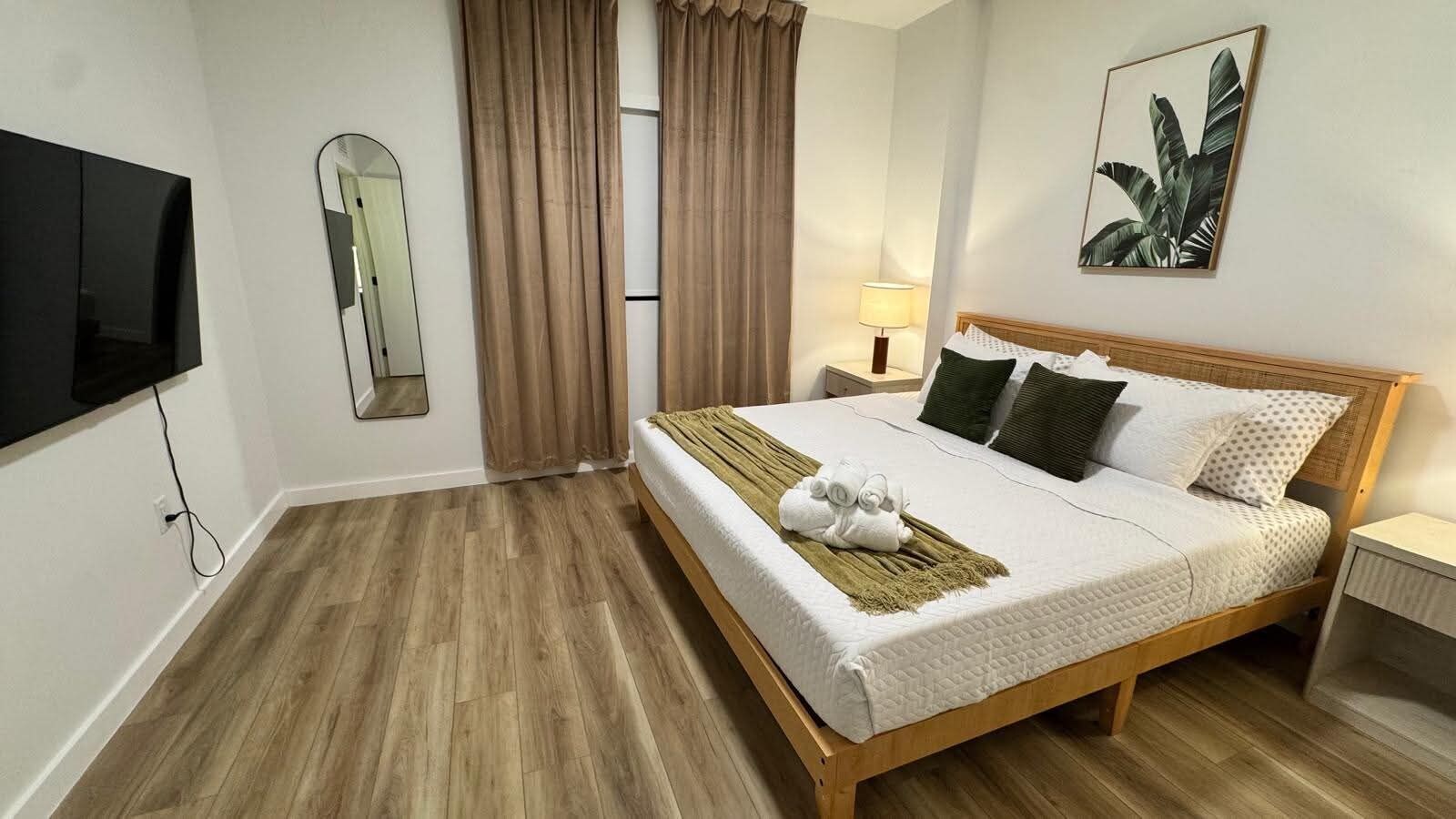 A hotel room with a wooden bed, a flat-screen TV mounted on the wall, a bedside table with a lamp, and a large window with curtains.