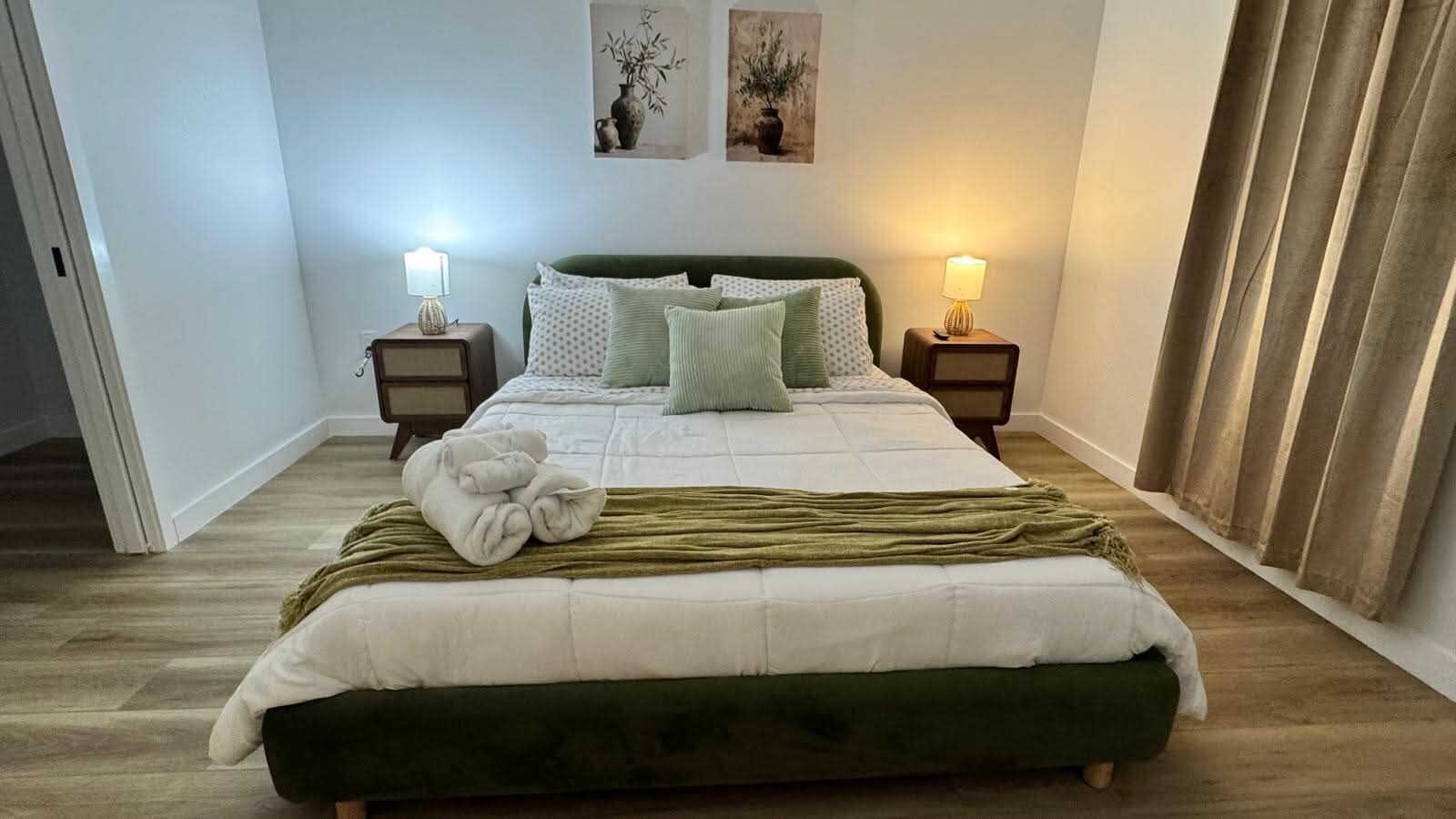 A neatly made bed with a green headboard, two bedside lamps, and a pair of framed botanical prints on the wall.