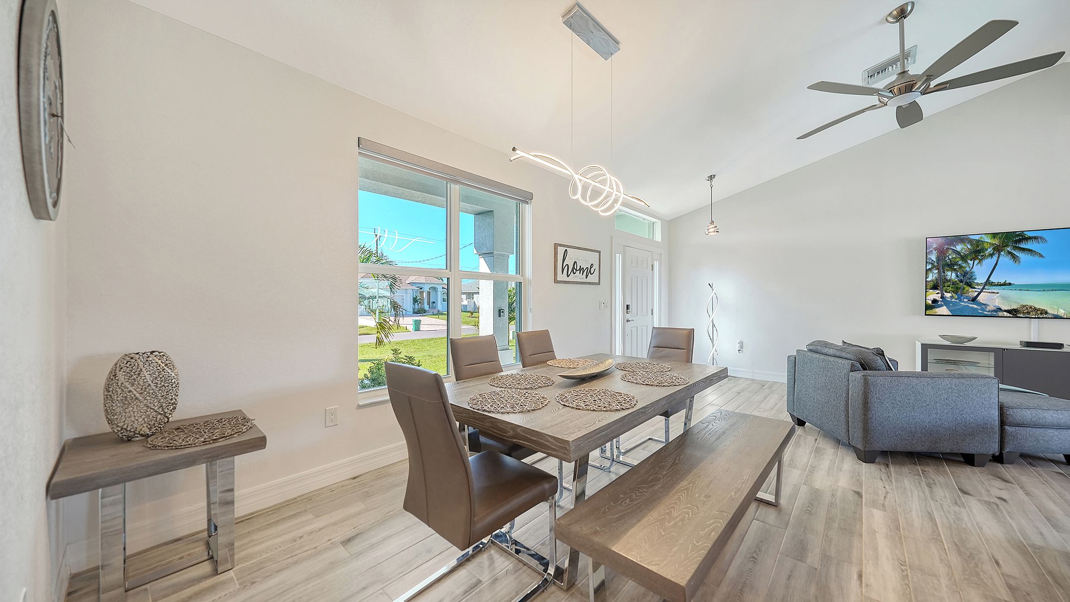 A modern living room with a dining area, a ceiling fan, and a television displaying a beach scene.