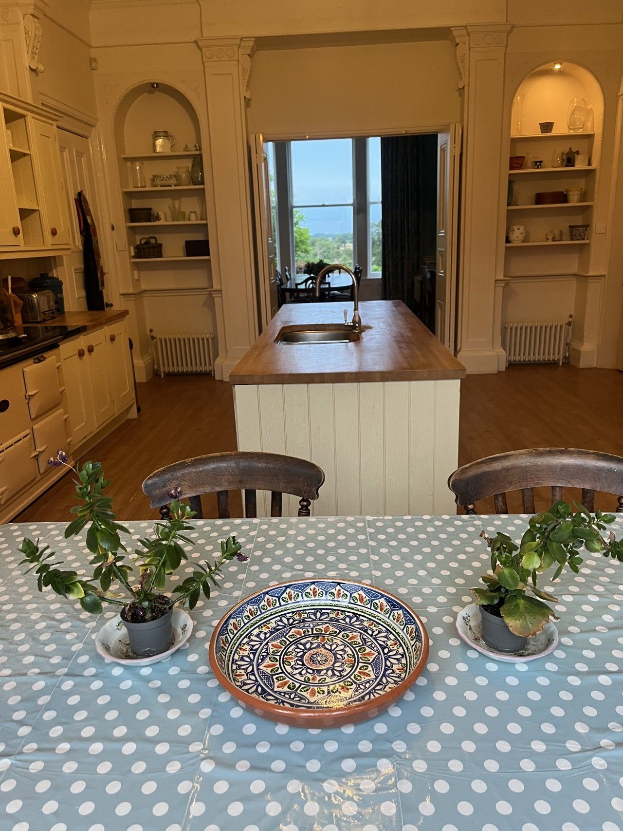 Un coin repas avec une table dressée pour deux personnes, agrémentée d’une nappe à pois bleus, de deux plantes en pot et d’une assiette décorative.
