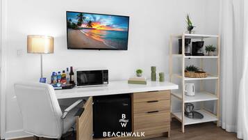 Una habitación moderna con un escritorio blanco, un televisor de pantalla plana, un microondas, una estantería y una pequeña planta.