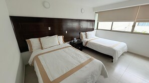 Habitación de hotel con dos camas, un cabecero de madera oscura, una mesita de noche con lámpara y una ventana con vistas al exterior.