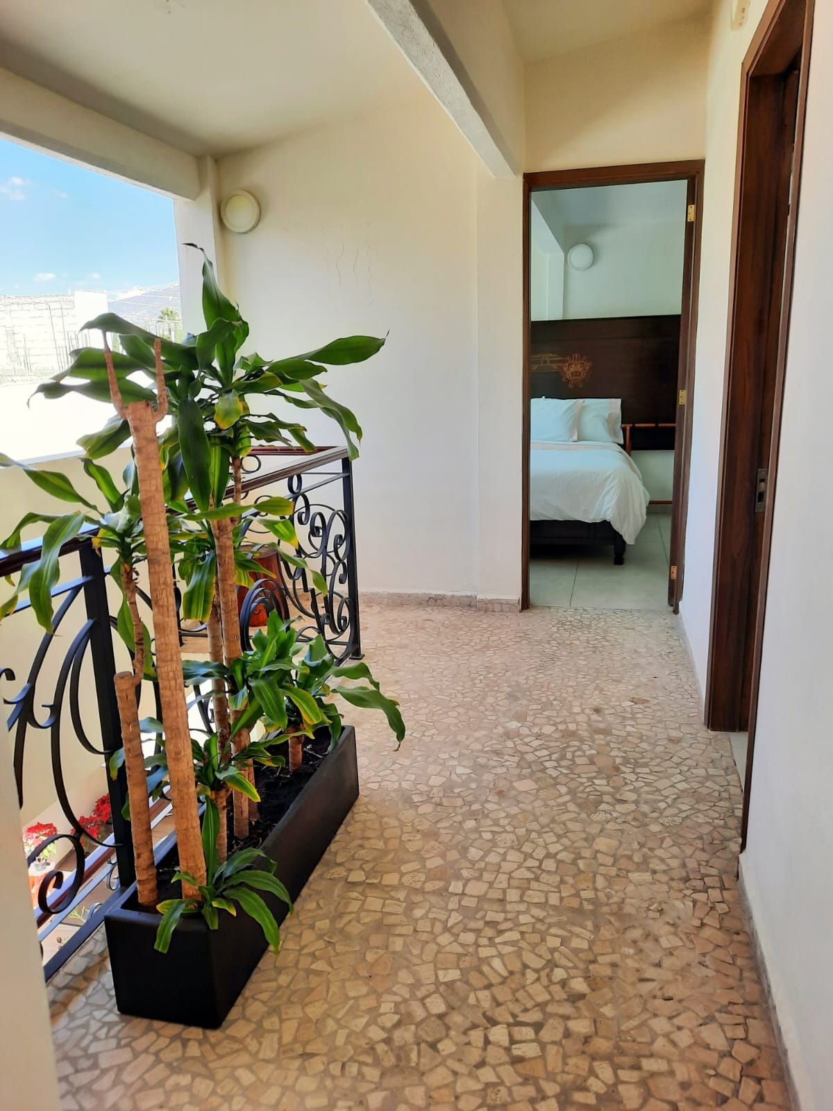 A narrow corridor with a tiled floor leading to a bedroom with a bed and a nightstand. There is a potted plant with a wooden stand on the left side.