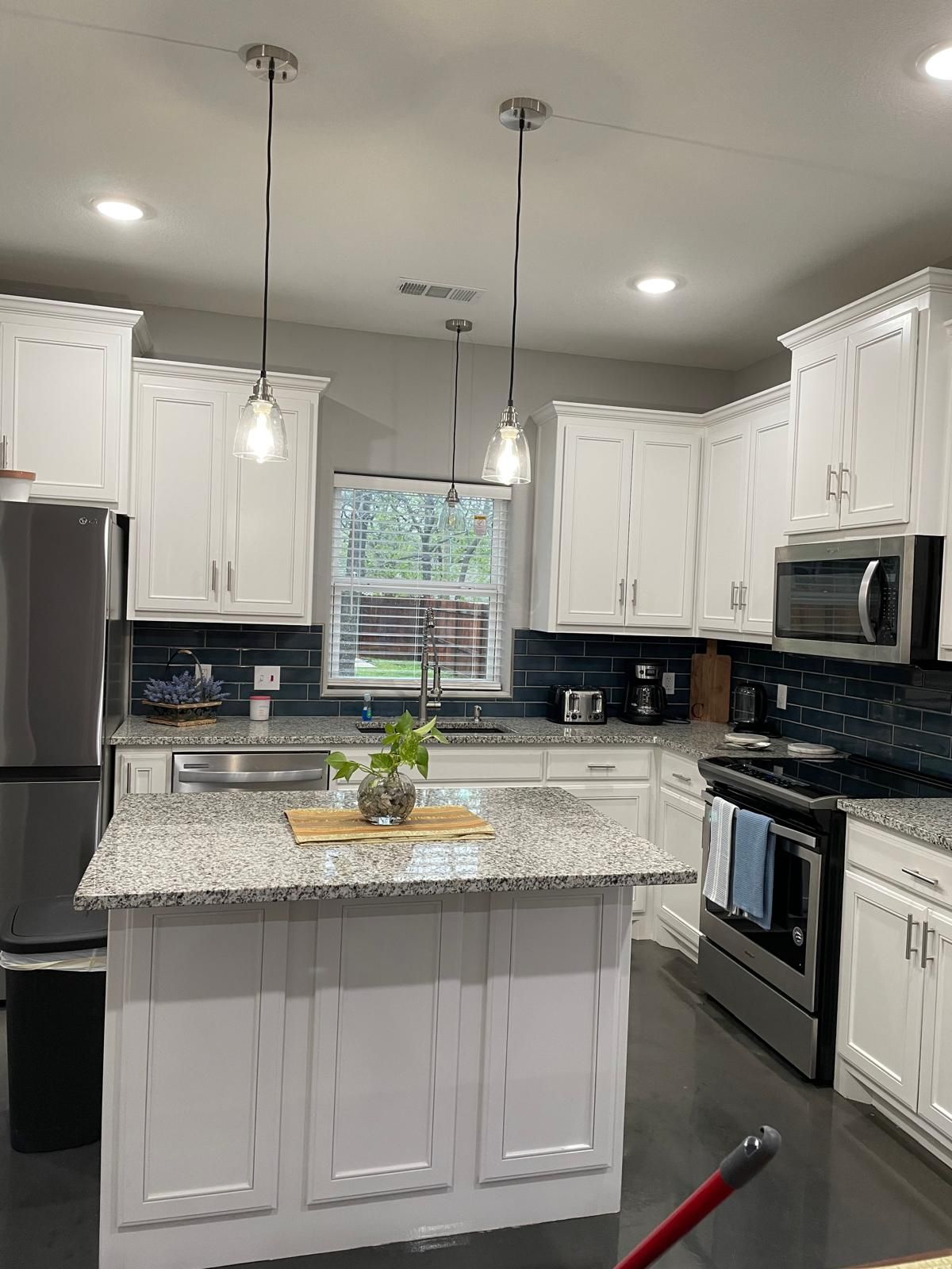 A modern kitchen with white cabinets, granite countertops, stainless steel appliances, pendant lighting, and a window with blinds.