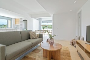 A modern living room with a sofa, a coffee table, and a TV. There is a dining area with chairs and a table. The room has large windows and a view of the outdoors. - Le Mont Blanc | 2-Bedroom Apartment with Shared Pool (Kounoupidiana)