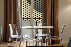 Un coin repas avec une table ronde et quatre chaises transparentes, une porte en verre ornée d’un motif géométrique et des rideaux marron.
