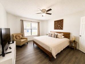 A bedroom with a bed, a chair, a television, a ceiling fan, a window with blinds, and a door. - Mid-Century Hideaway - Near SeaWorld (San Antonio)