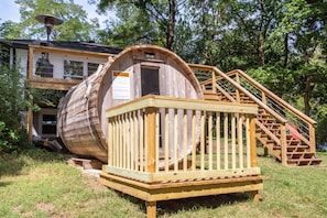 Een houten vatstructuur met een houten trap naar een deur, omringd door gras en bomen.