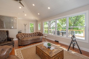 A spacious living room with a large window, a brown leather sofa, a wooden coffee table, and a tripod.