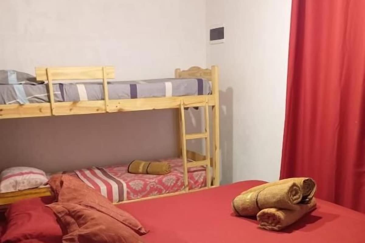 A room with a bunk bed, a single bed with red bedding, a red curtain, and a tray with a phone and a black object.