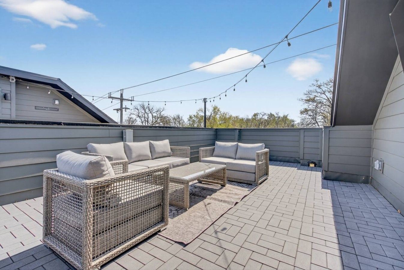 An outdoor patio with a sofa set and a wicker chair, a brick floor, and overhead string lights.