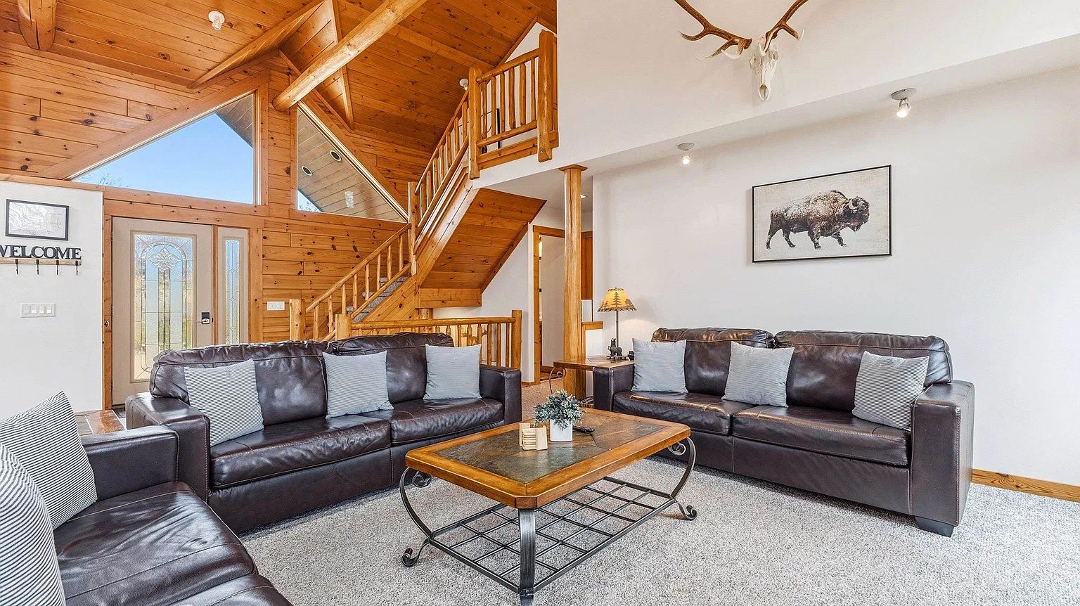 A spacious living room with a wooden staircase, leather sofas, and a mounted buffalo picture.