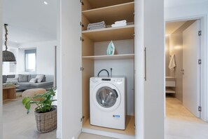 A modern laundry room with a front-loading washing machine, a drying rack, and neatly folded towels on shelves. - Le Mont Blanc | 3-Bedroom Apartment with Shared Pool (Kounoupidiana)