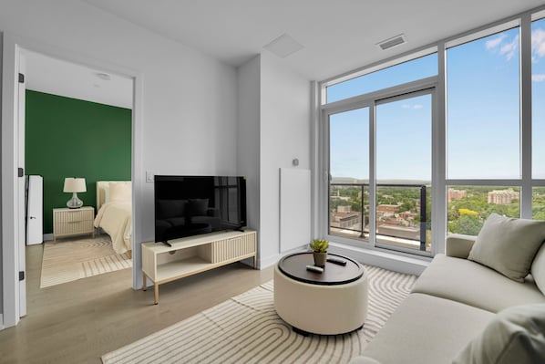 A modern living room with a large TV, a sofa, a round coffee table, and a view of a cityscape through large windows.