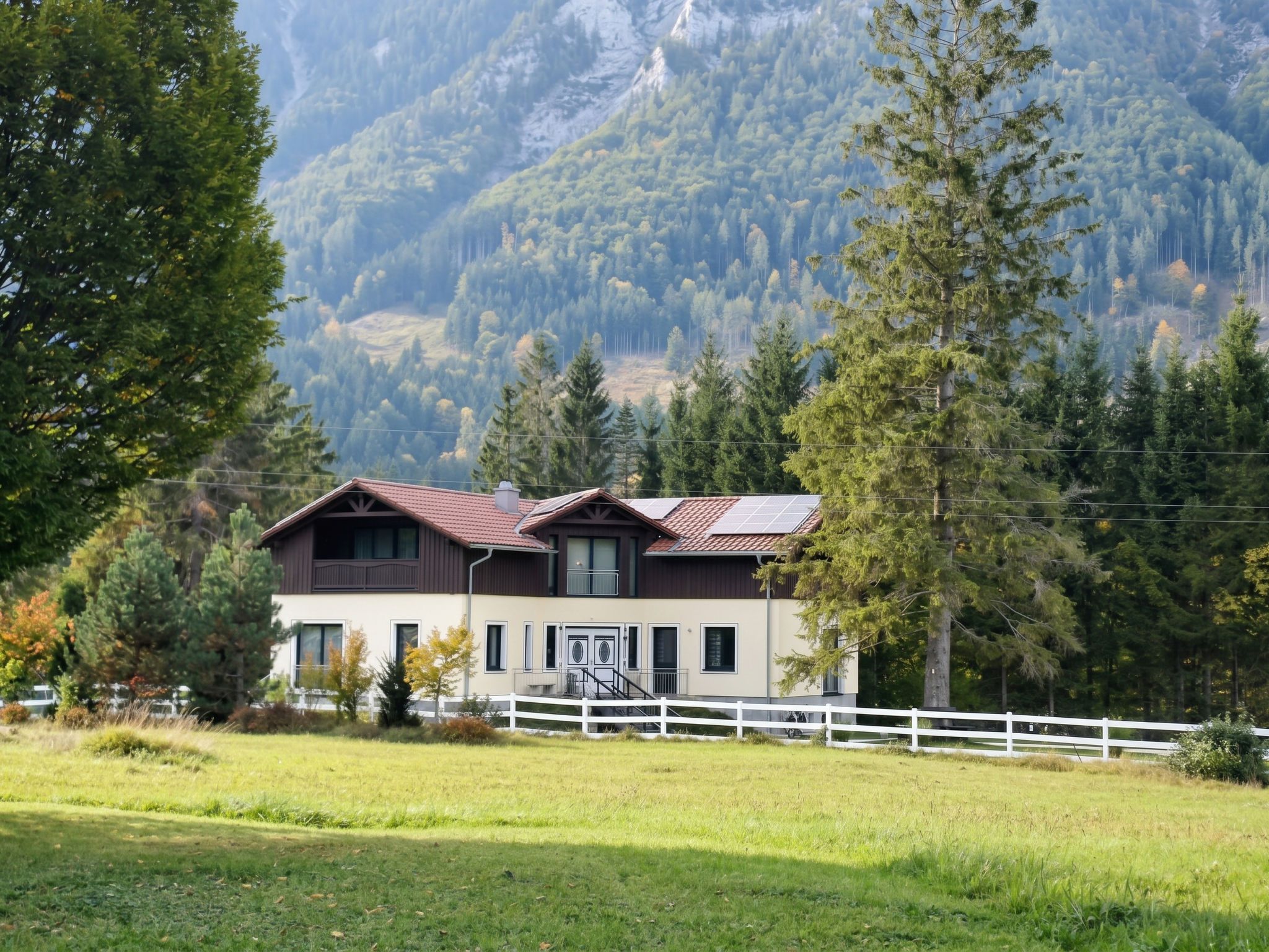 Een huis met twee verdiepingen, een schuin dak, een witte hekken en een bergachtige achtergrond.