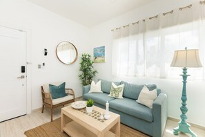 A living room with a sofa, a coffee table, a chair, a lamp, a mirror, and a window with curtains. - Apartment with a Pool Just Steps from the Beach and Restaurants, Central Location (Punta Cana)
