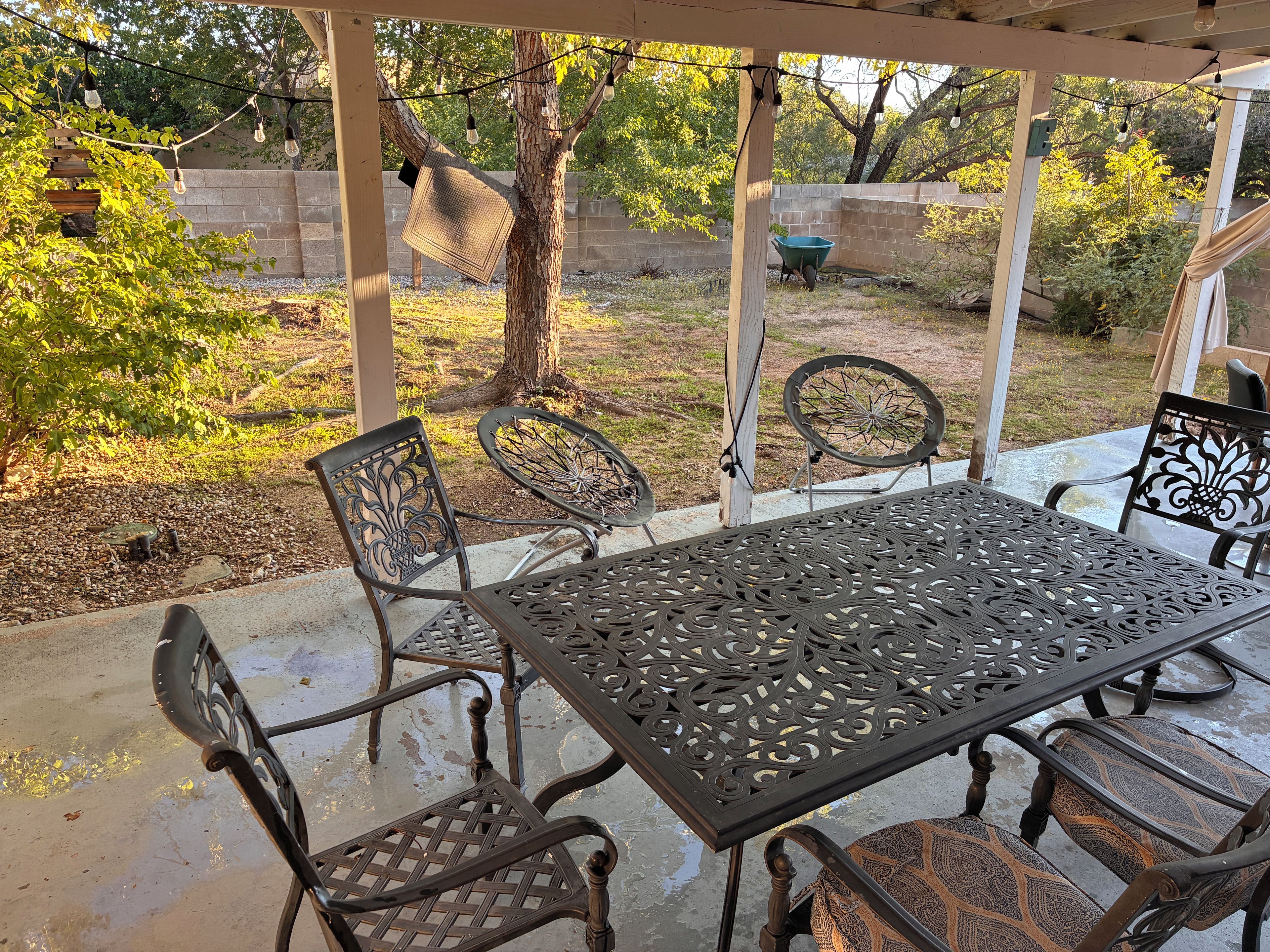 An outdoor patio with a metal table and chairs, surrounded by a wooden pergola and string lights.