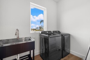 A laundry room with a sink, two washing machines, and a window with a view of the sky and trees. - Vilano Villa | Pool | Wellness | Water View | Golf (St. Augustine)
