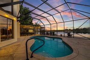 An indoor swimming pool with a view of a sunset over a body of water, surrounded by a covered patio area with seating. - Lakefront Estate 16ppl |Pool| Sauna| Dock| Hot Tub (Sebring)