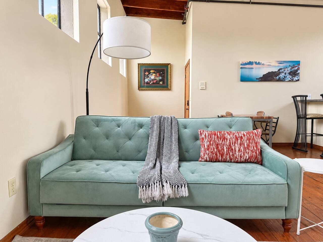 A teal sofa with a patterned throw blanket and a red pillow, a white table with a blue vase, and a painting of a coastal scene on the wall.