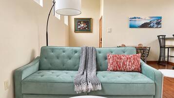 A teal sofa with a patterned throw blanket and a red pillow, a white table with a blue vase, and a painting of a coastal scene on the wall.