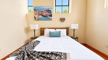 A hotel room with a large bed, two bedside tables with lamps, a view of a coastal town through a window, and a wall-mounted picture.