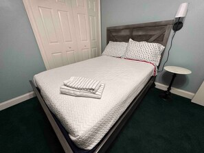 A neatly made bed with a white quilt and folded towels, a bedside table with a lamp, and a door in the background.