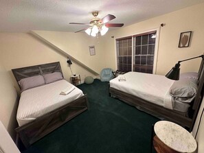 A bedroom with two beds, a ceiling fan, a window with blinds, a bedside lamp, and a small round table.
