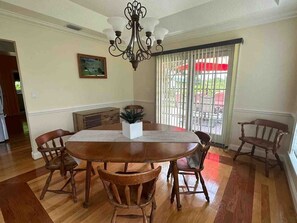 A dining room with a round table, six wooden chairs, a chandelier, a potted plant, and a window with blinds.