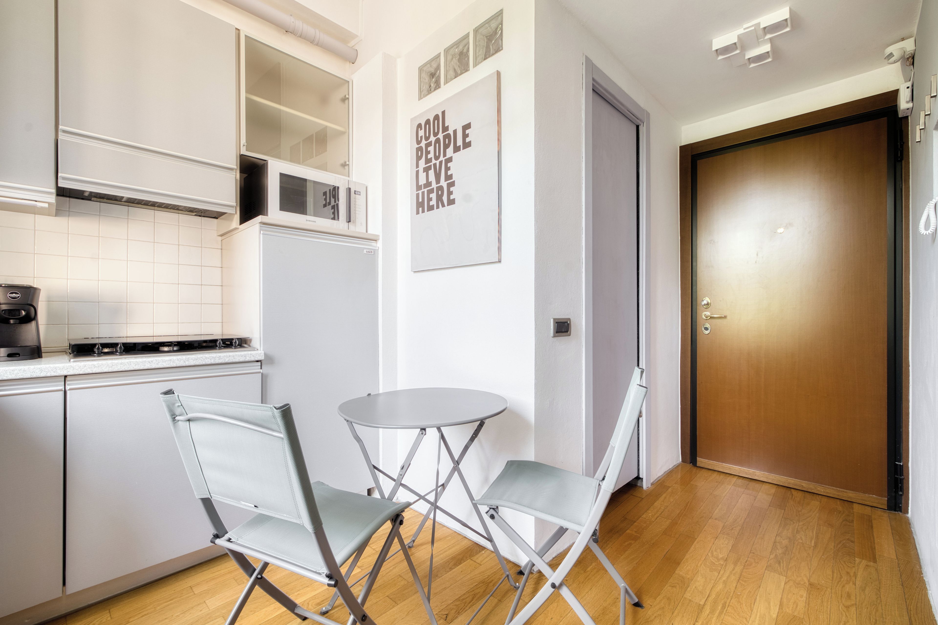 A compact kitchen with white cabinets, a stainless steel range hood, a microwave, and a small dining table with two chairs.