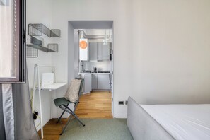Habitación pequeña con cama, escritorio y silla. Se ve una kitchenette al fondo.