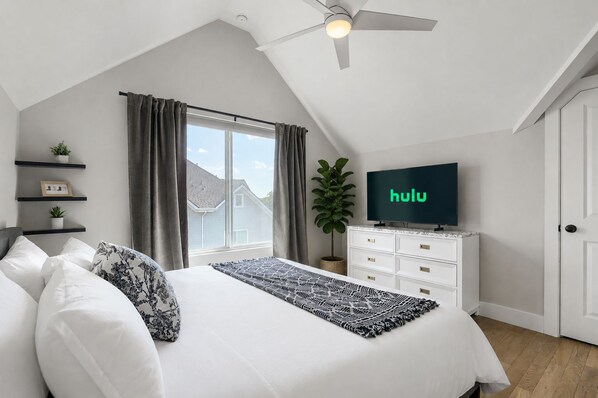 Un dormitorio con cama, televisión, ventilador de techo, ventana con cortinas y una planta.
