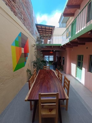 Un comedor con una larga mesa de madera y sillas, un mural geométrico colorido en la pared y un balcón con barandilla verde.