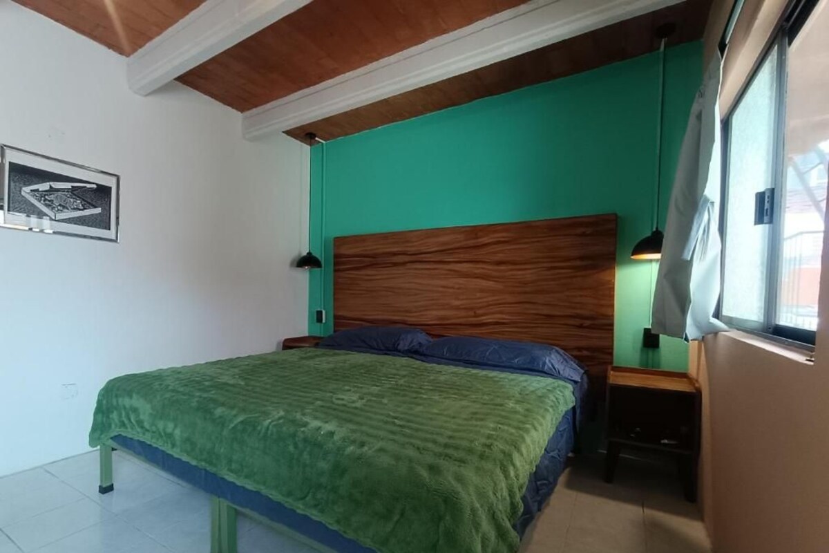 Un dormitorio con una cama, cabecera de madera, pared verde, ventana y mesita de noche con una lámpara.
