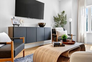 Een moderne woonkamer met een flatscreen televisie, een grijze bank, een houten salontafel en een potplant.