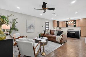 A modern living room with a sofa, coffee table, and chairs, a kitchen area with wooden cabinets, and a ceiling fan. - Aboretum. White Rock Lake. Modern 3 Br. 6 guests (Dallas)
