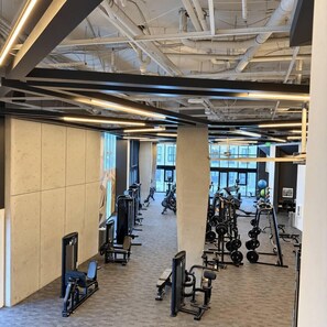 Ein Fitnessraum mit Trainingsgeräten, großen Fenstern und einer hohen Decke mit freiliegenden Lüftungsrohren.